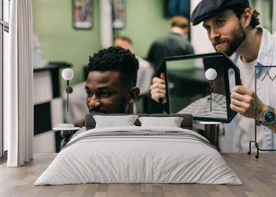 Portrait of handsome black man with comb in hair looking in the mirror at his new haircut. Barber hairdresser showing client his work. Male beauty treatment concept. Pretty young African guy face Wall mural